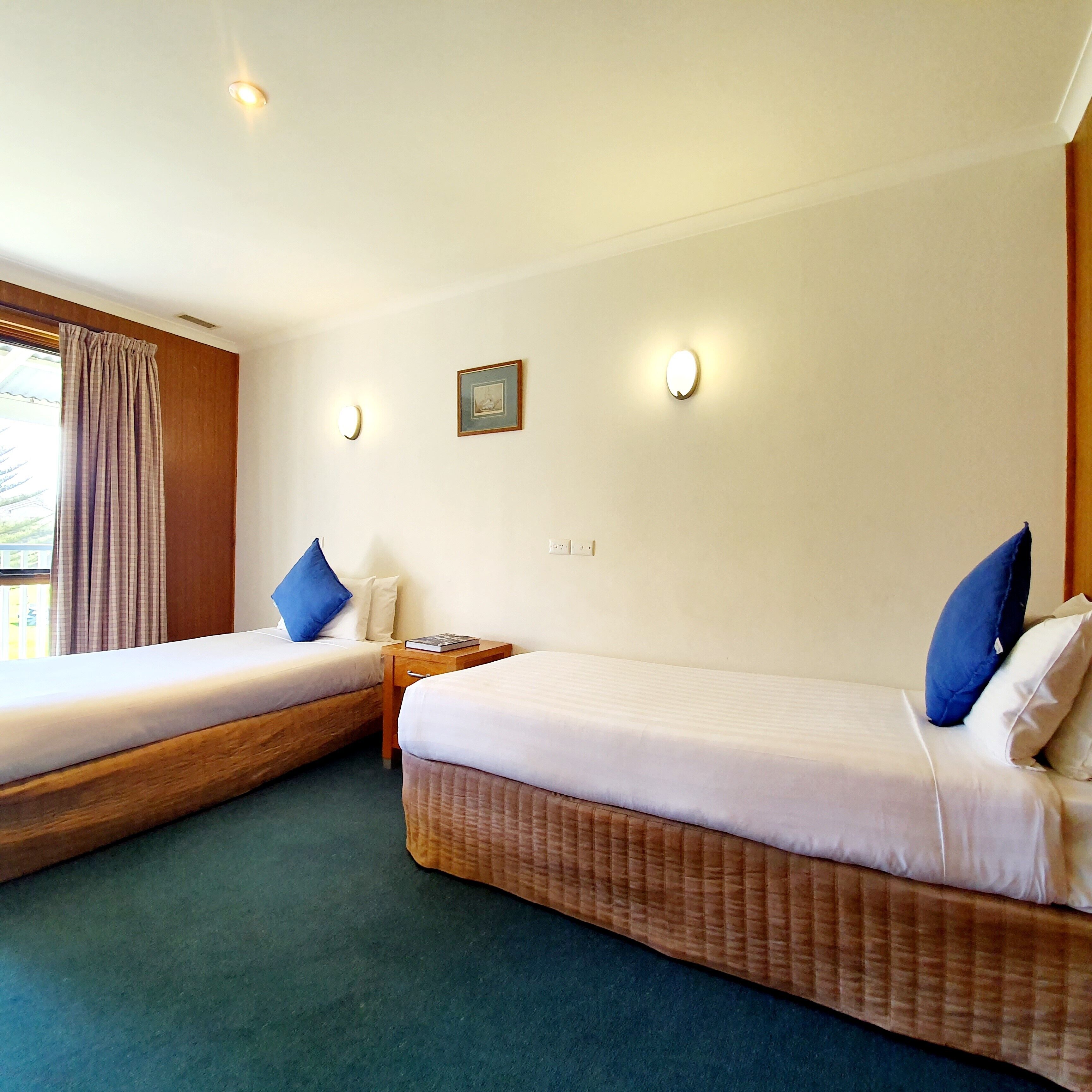 Habitación de hotel con dos camas individuales, cabecero de madera, mesita de noche y un cuadro enmarcado en la pared, además de una ventana con cortinas.
