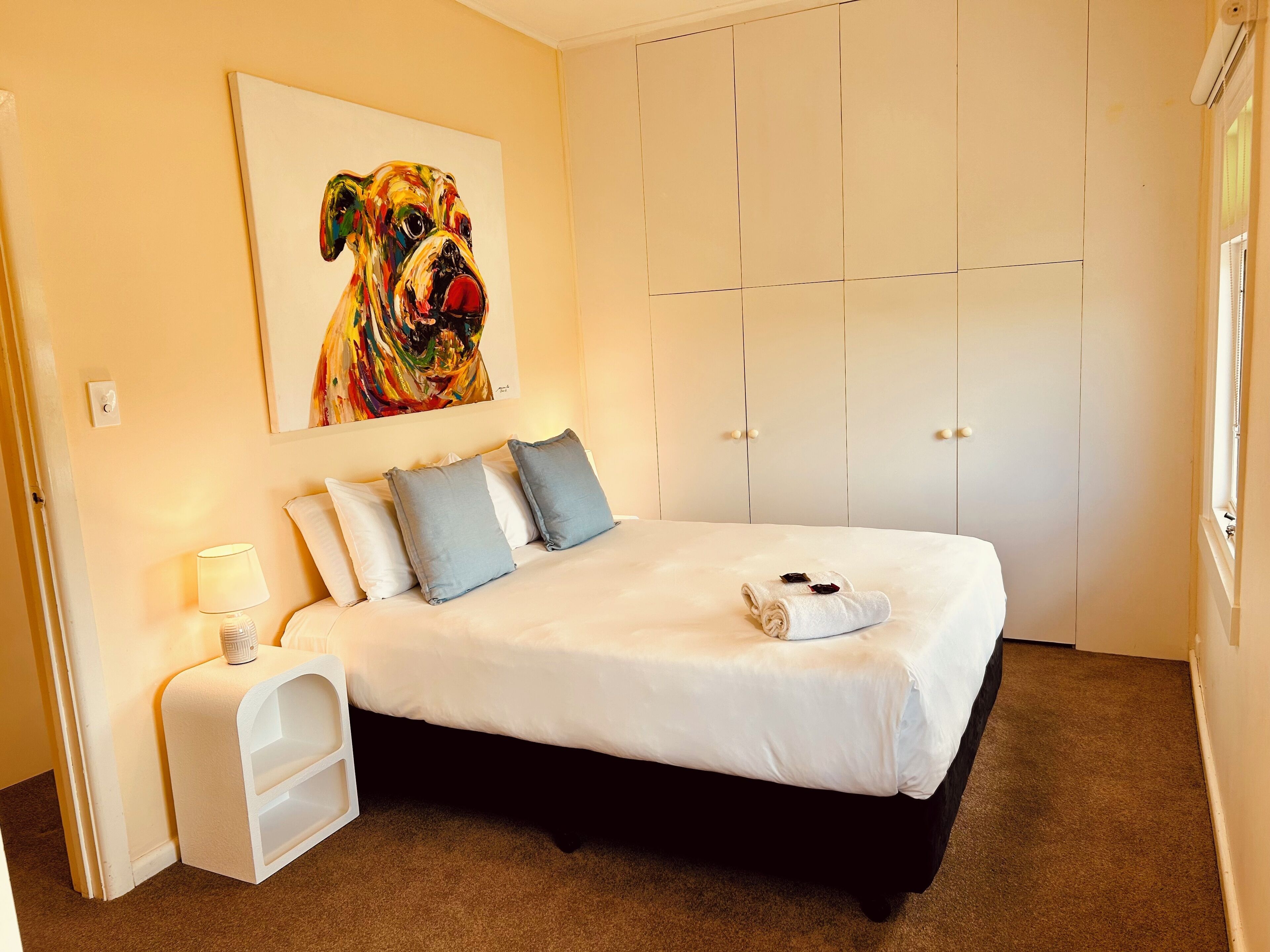 Een slaapkamer met een bed, kussens, een nachtkastje, een lamp, een schilderij en een kledingkast.