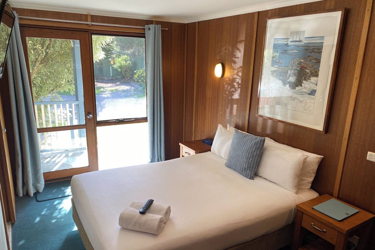 A hotel room with a large bed, a nightstand, a framed picture on the wall, and a view of the outdoors through the window.