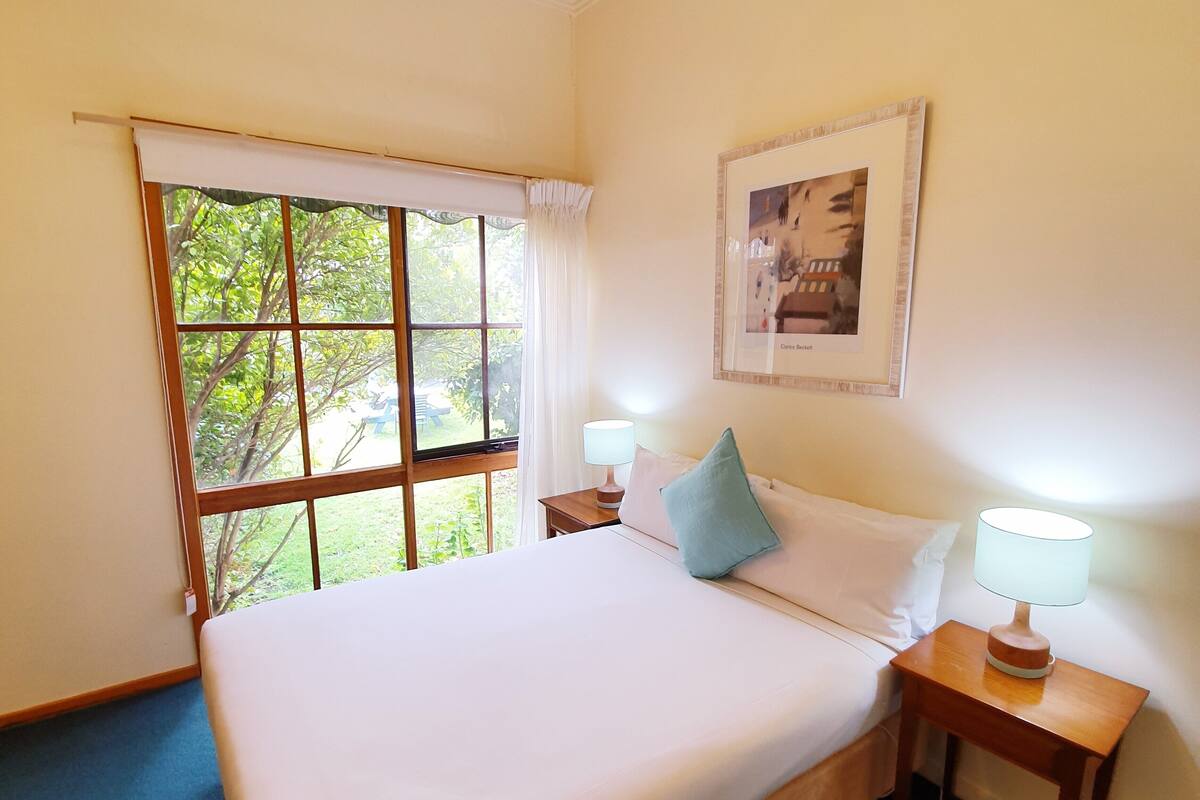 A neatly made bed with white linens, a window with a view of greenery, and a framed picture on the wall.