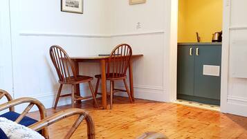 A room with a wooden floor, a dining table with chairs, a bed with a blue mattress, and a kitchen area with a yellow wall and cabinets.