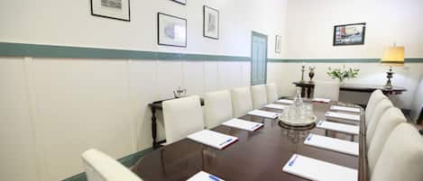 Un salón de conferencias con una larga mesa, sillas blancas, cuadros en la pared y una lámpara a un lado.