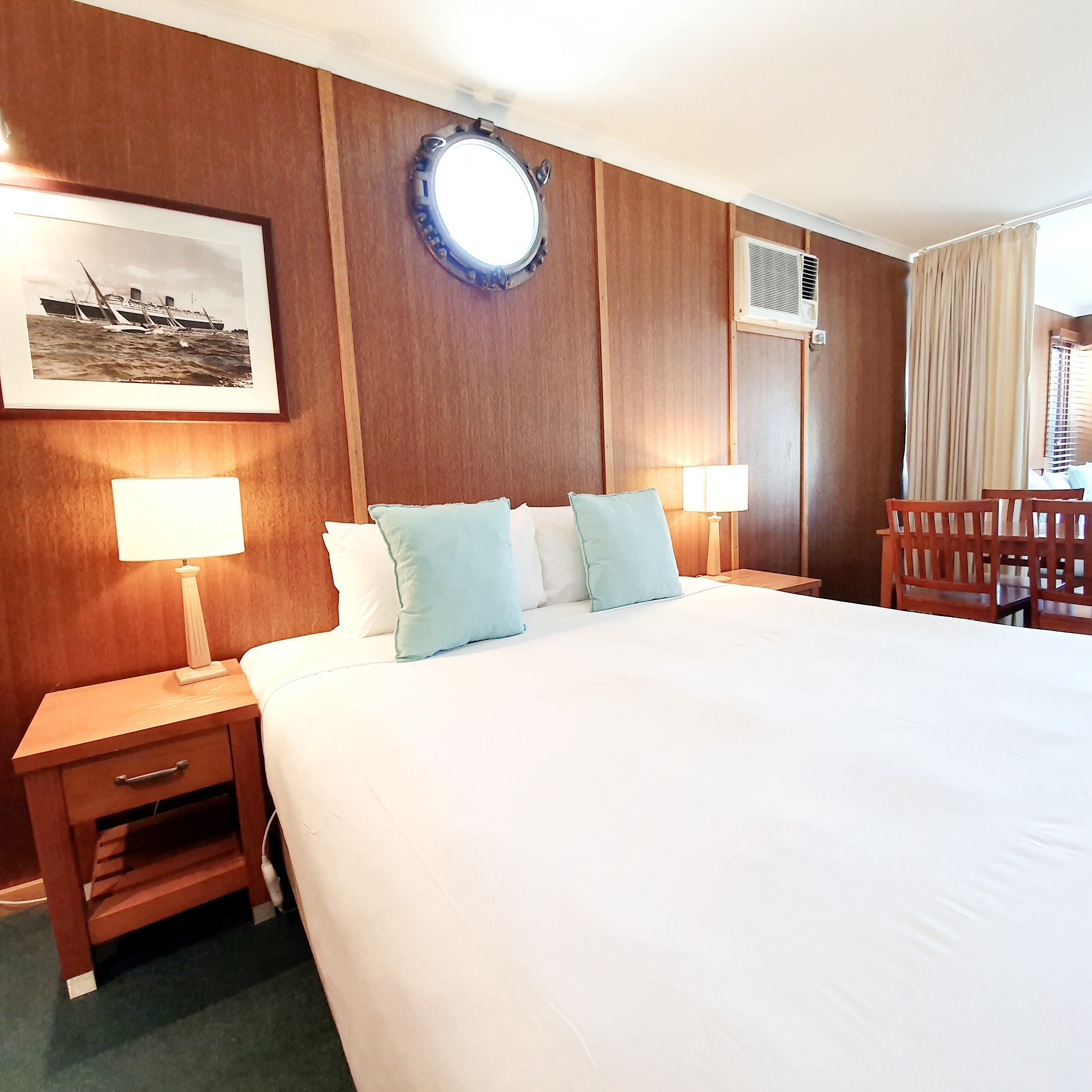 Ein Hotelzimmer mit einem großen Bett, zwei Nachttischen mit Lampen, einem Kopfteil, einer Wand-Klimaanlage, einem Fenster mit Vorhängen und einem gerahmten Bild an der Wand.