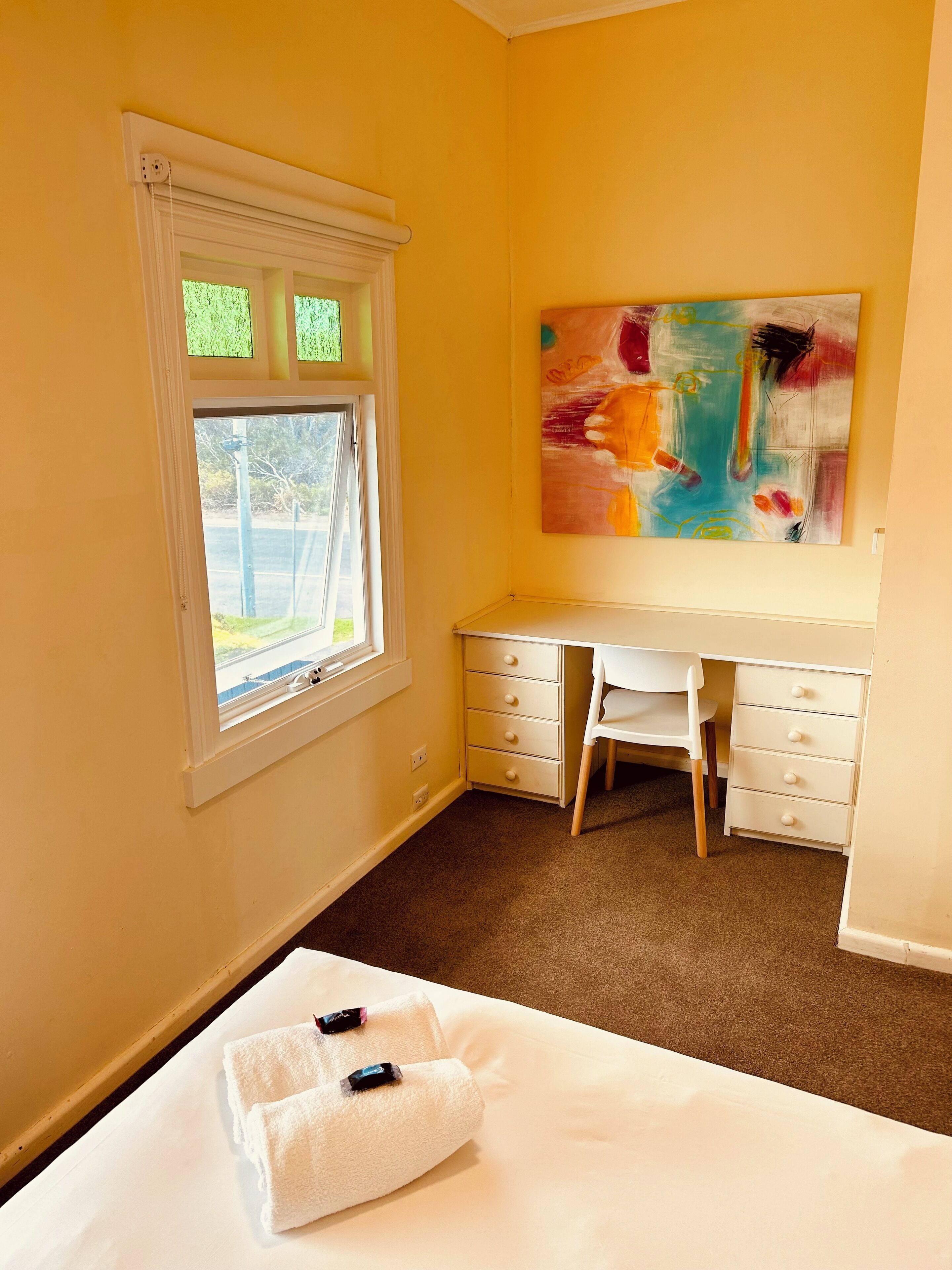 Un dormitorio con cama, escritorio, silla, ventana y un cuadro en la pared.