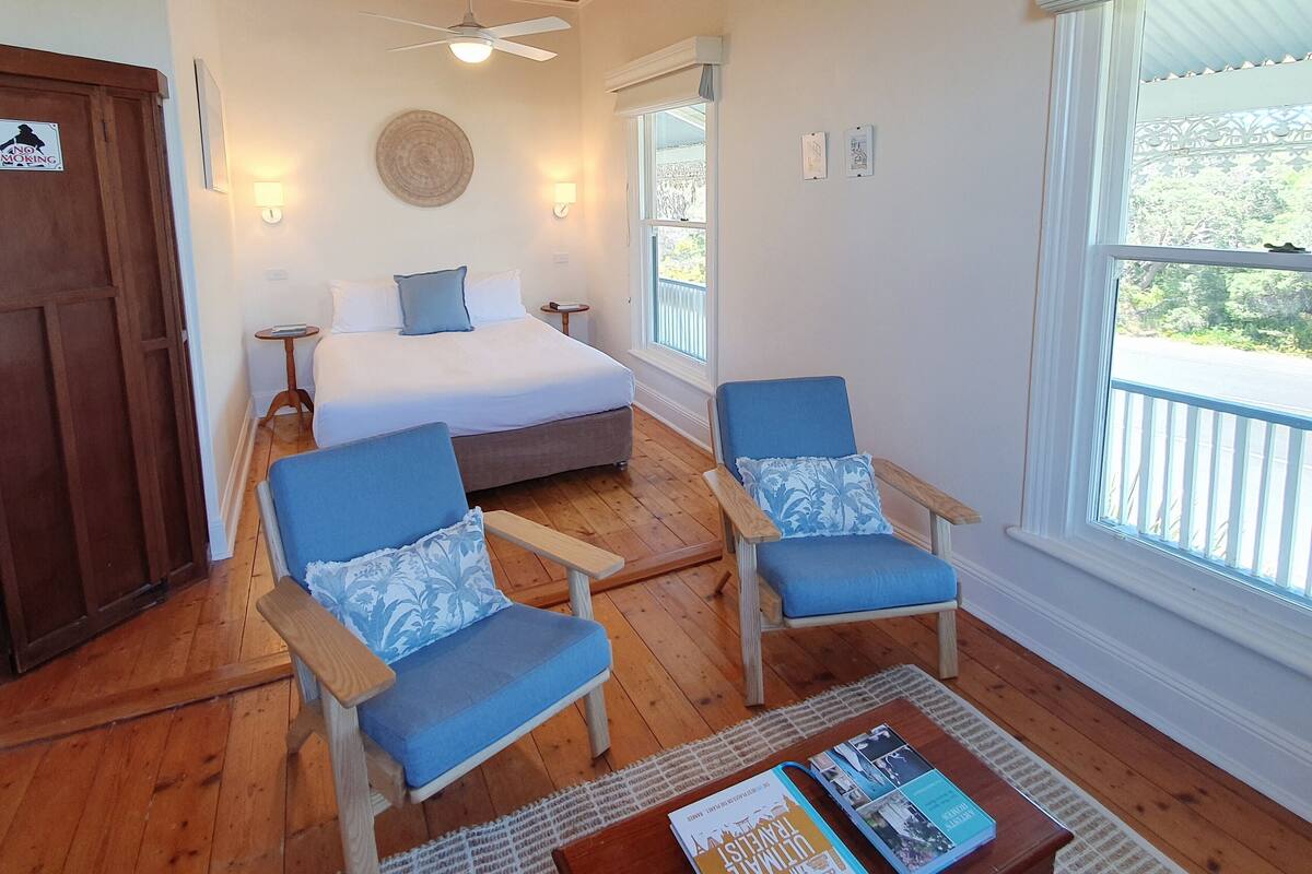 A bedroom with a bed, two chairs, a ceiling fan, a window, and a nightstand.