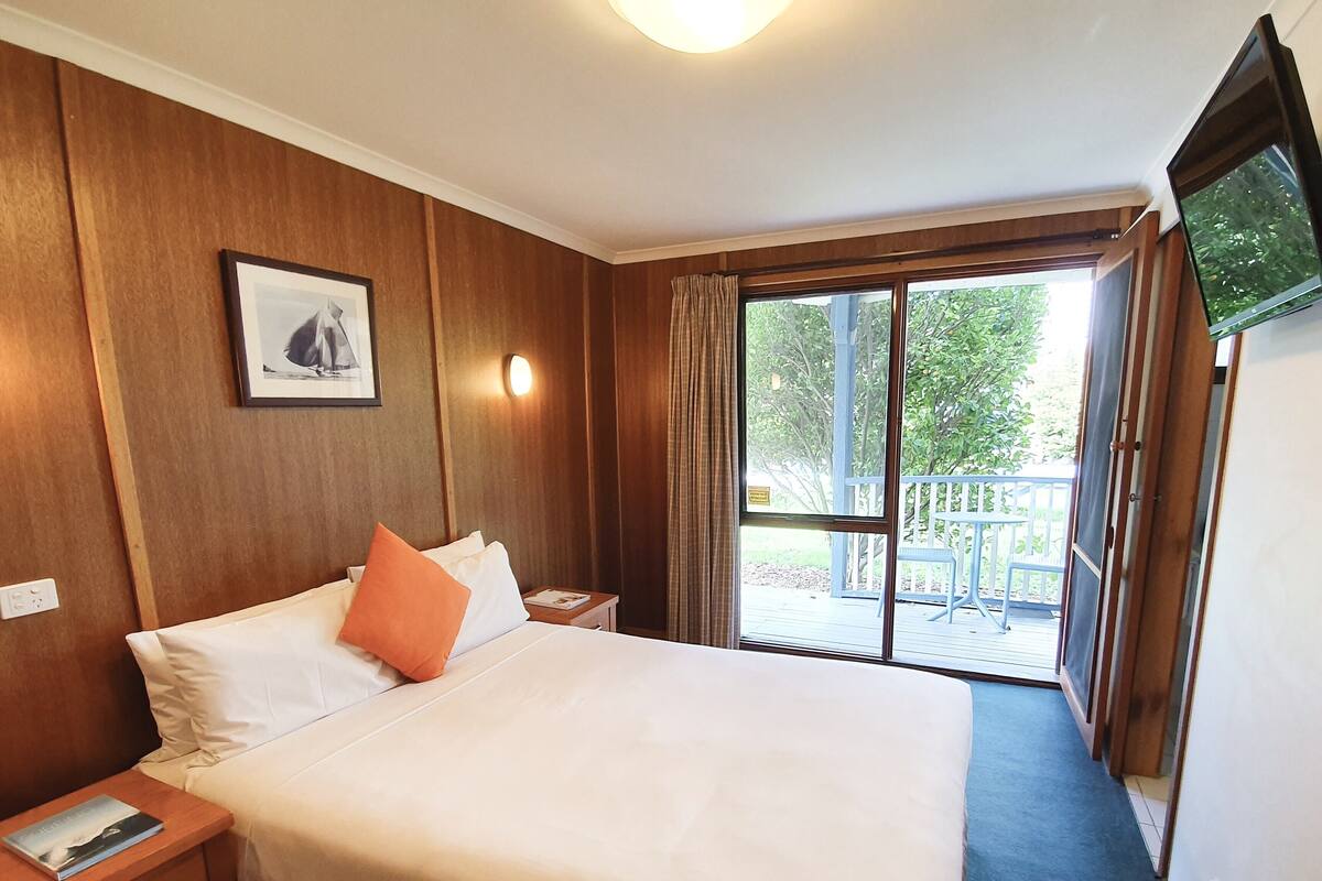 A hotel room with a large bed, a nightstand, a framed picture, a ceiling light, and a view of the outdoors through a sliding door.