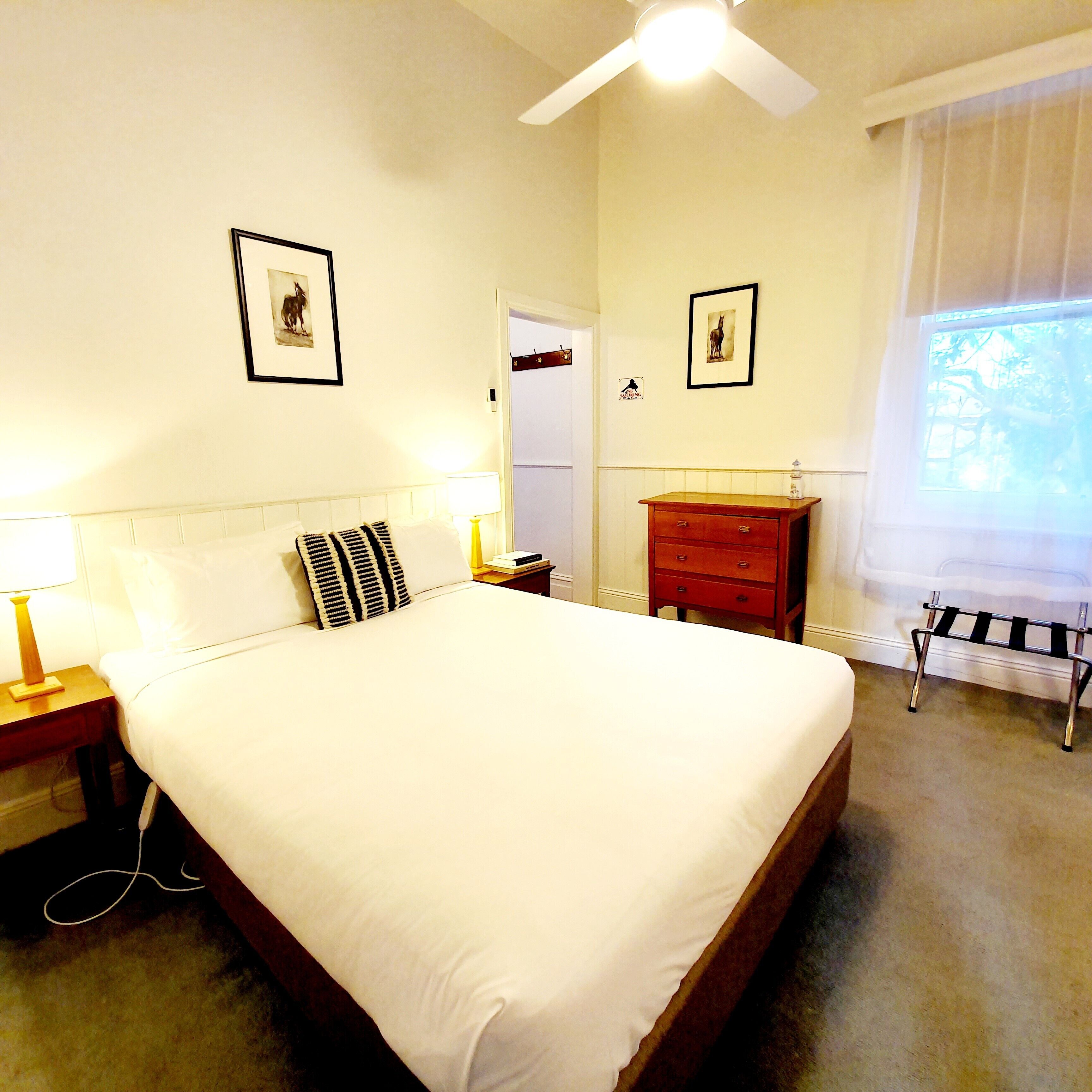 A neatly made bed with white bedding, a wooden nightstand with a lamp, a red dresser, and two framed pictures on the wall.