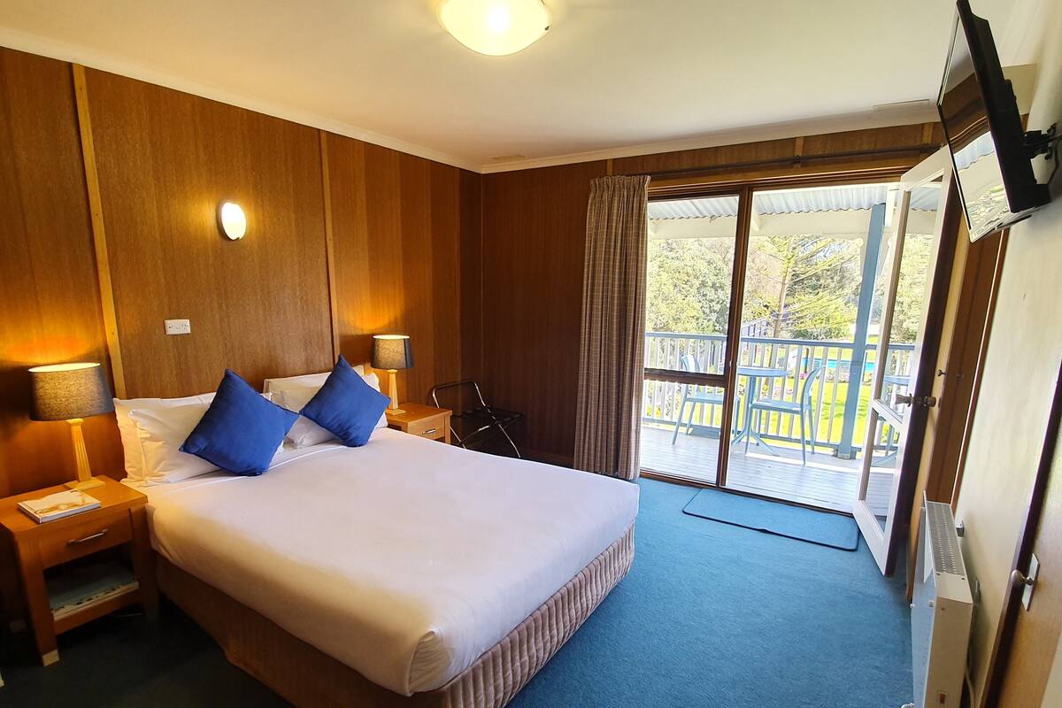 A hotel room with a large bed, a nightstand, a lamp, a window with curtains, and a view of the outside deck.