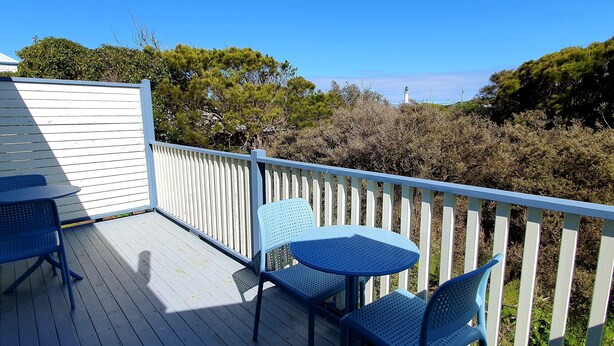 A wooden deck with two blue chairs and a table, surrounded by a white railing, overlooking a landscape with trees and a lighthouse in the distance. - Point Lonsdale Guest House (Point Lonsdale)