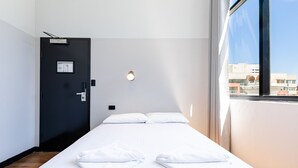 A hotel room with a bed, a door, a light fixture, and a window with a view of buildings. - Bunk Backpackers (Fortitude Valley)