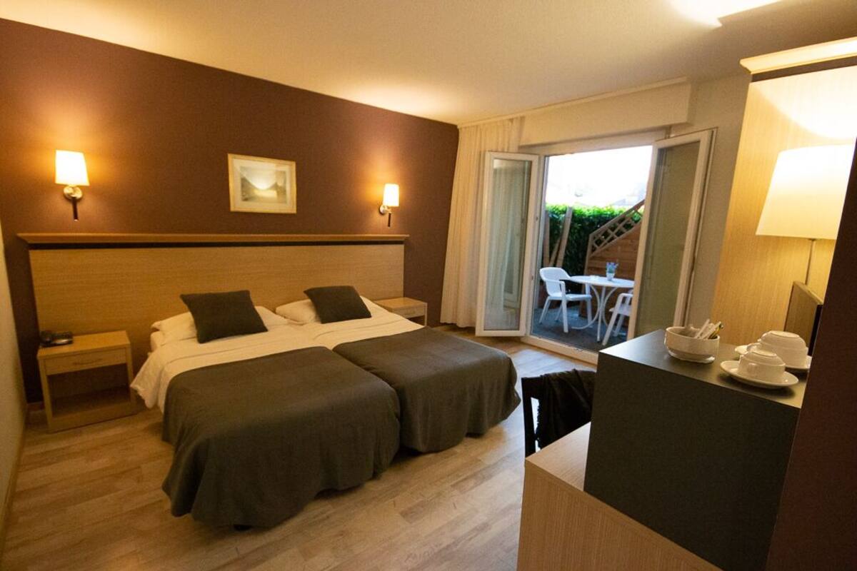 A hotel room with two beds, a desk, and a chair. There is a window with a view of greenery outside.