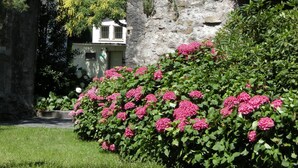 Un arc en pierre ancien, avec un bâtiment en arrière-plan et des fleurs roses au premier plan.