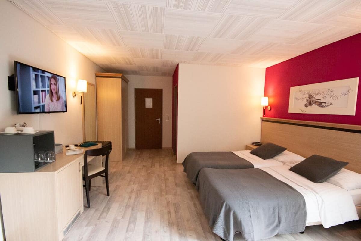 A hotel room with two beds, a desk with a chair, a TV, and a red accent wall.