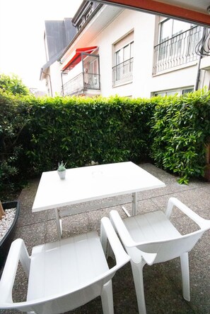 Uma mesa branca e duas cadeiras brancas numa varanda exterior, com um viveiro e um edifício ao fundo.