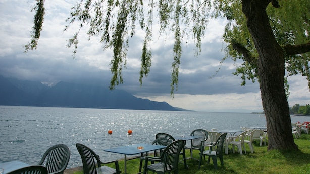 Un espace aménagé avec des tables et des chaises, situé au bord du lac, d’où l’on peut admirer le paysage et les montagnes au loin.