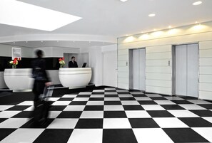 Zona de recepción de un hotel con suelo a cuadros, mostradores y ascensores.
