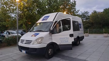 A white van parked in an outdoor area with a paved floor, surrounded by trees and a street lamp.