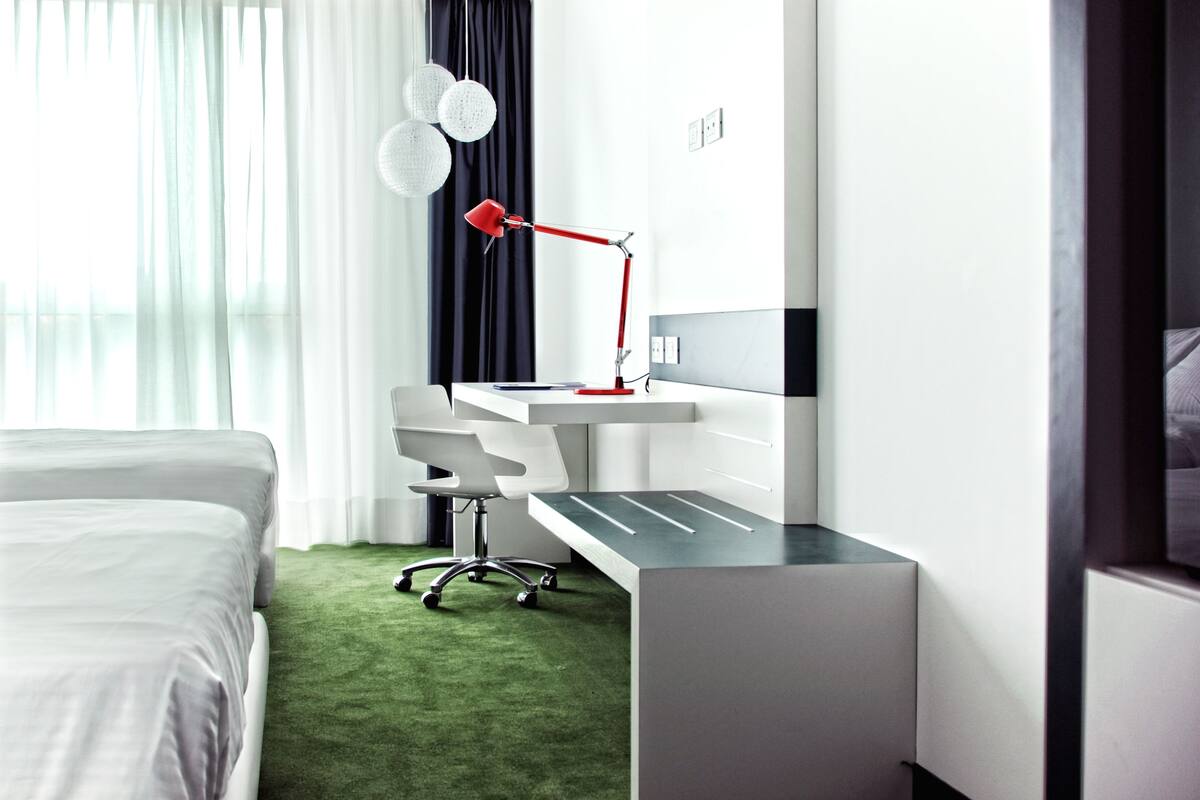 A modern hotel room with a neatly made bed, a desk with a red lamp, and a green carpet.