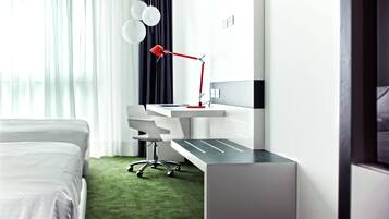 A modern hotel room with a neatly made bed, a desk with a red lamp, and a green carpet.