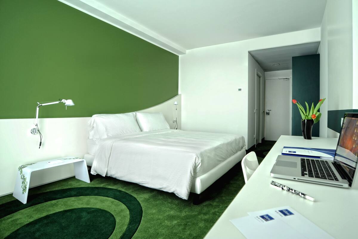 A hotel room with a bed, a desk with a laptop, a bedside lamp, and a small table with a plant.