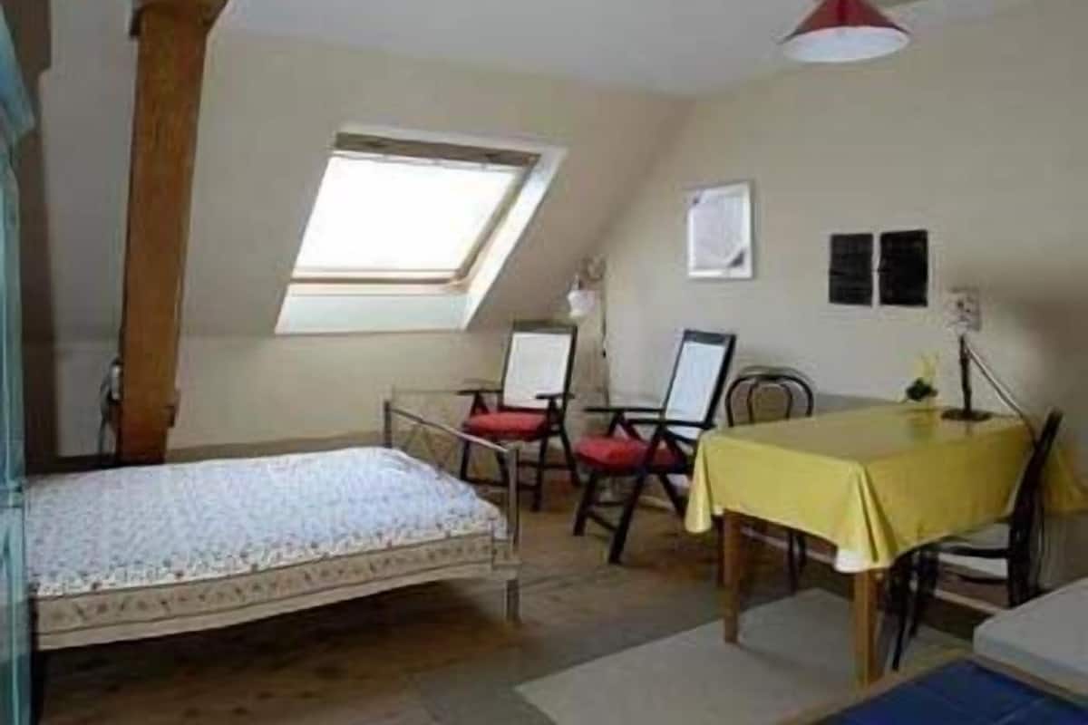 Una habitación pequeña y acogedora con una cama individual, una mesa y dos sillas. Tiene un tragaluz y un estante pequeño sujeto a la pared.