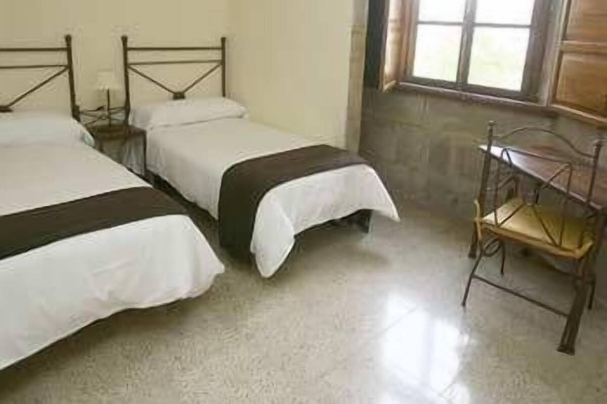 Una habitación con dos camas individuales, una silla de madera y un ventanal con cortinas.