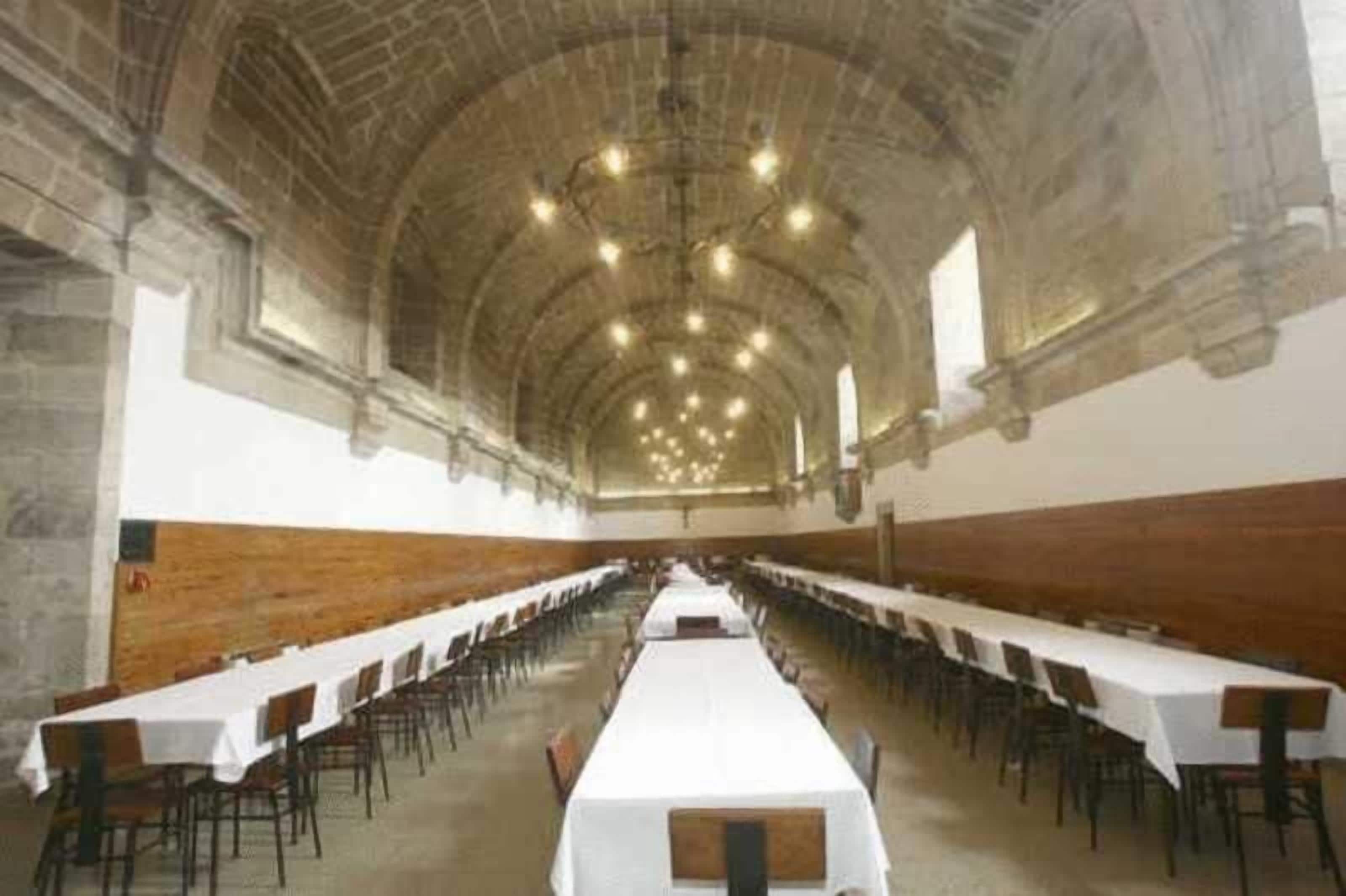 Een lange hal met rijen tafels en stoelen, een gewelfde plafond en stenen muren.