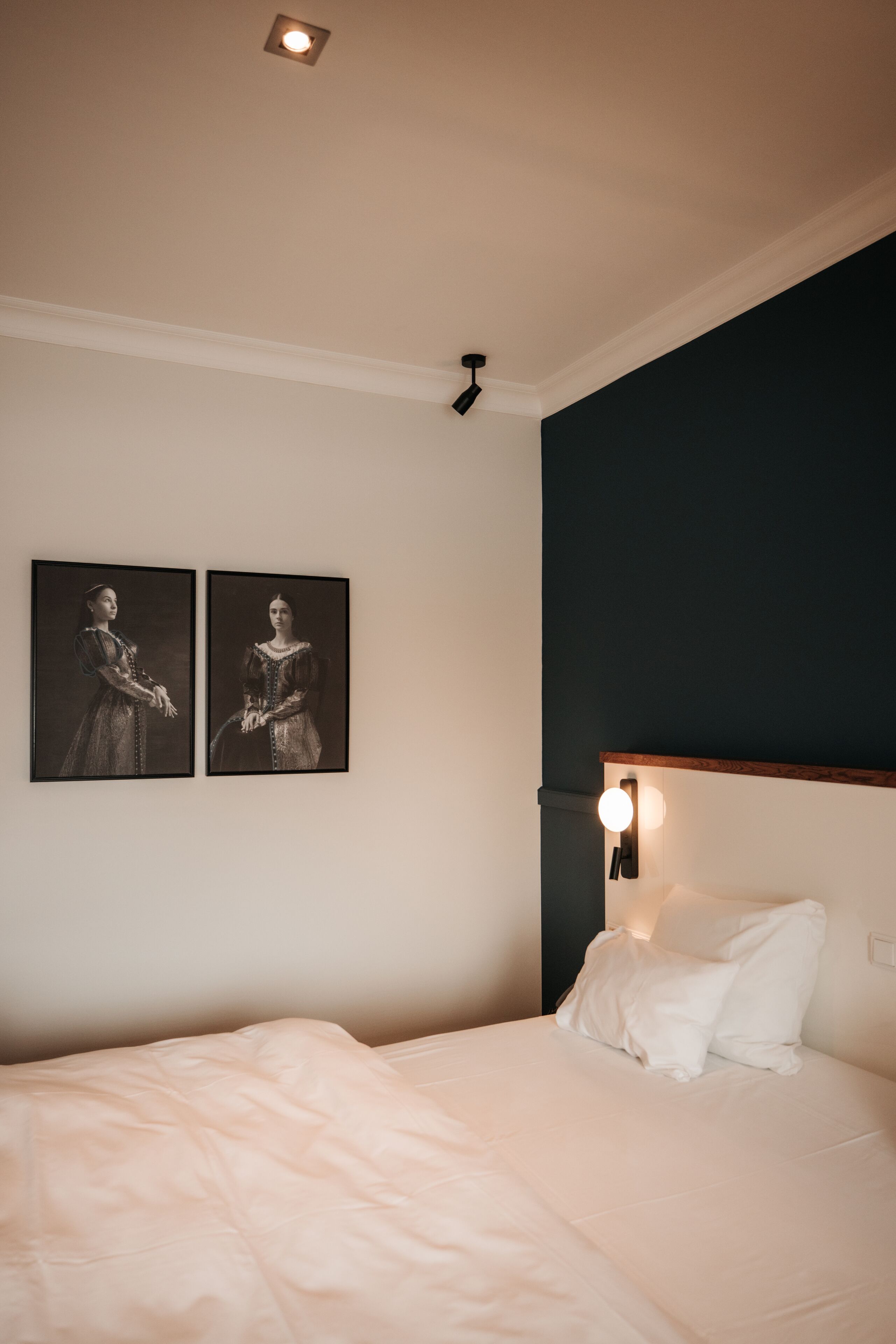 Ein Schlafzimmer mit einem Bett, zwei gerahmten Portraits an der Wand, einer Deckenleuchte und einer Nachttischlampe.