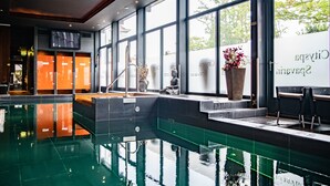 A modern indoor swimming pool with clear glass walls, a seating area with a potted plant, and a staircase leading to an upper level.