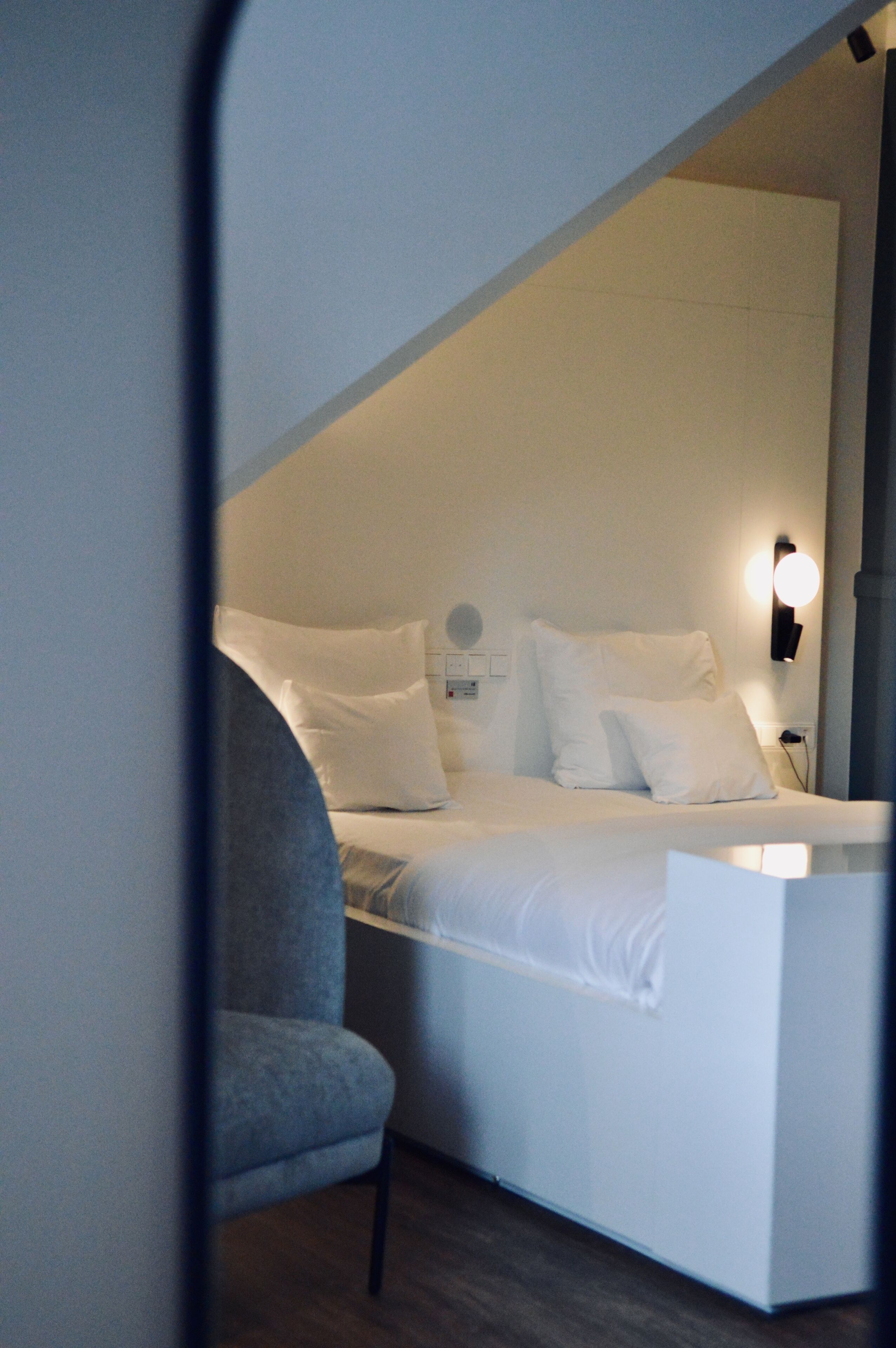 A bedroom with a bed, pillows, a chair, and a wall-mounted light fixture.