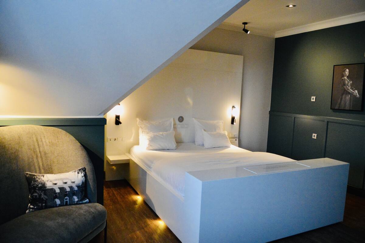 A modern bedroom with a large bed, a nightstand, a chair, and a painting on the wall.