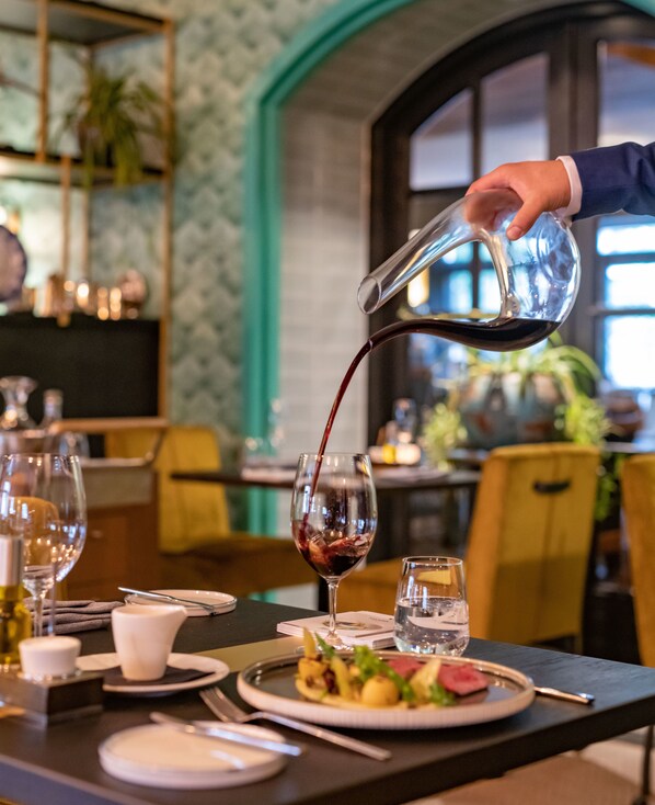 A dining table with a plate of food, a glass of red wine, and a hand pouring wine.