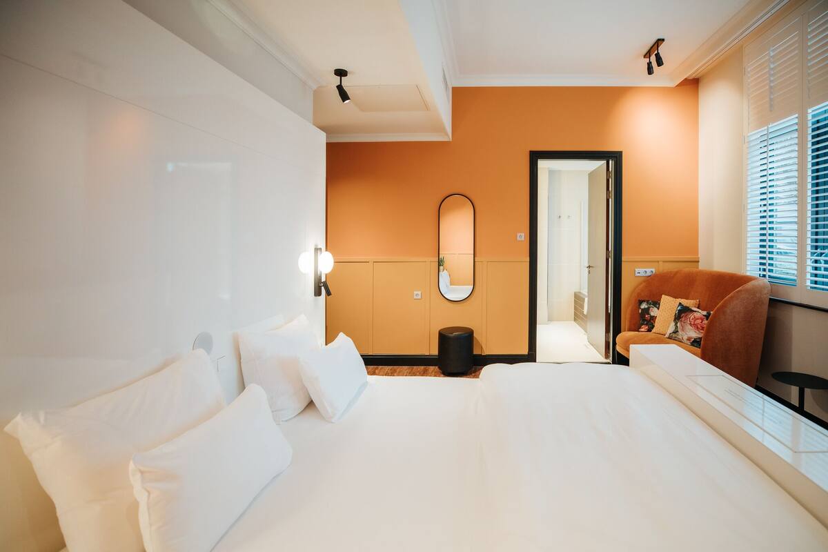 A hotel room with a large bed, a sofa, a mirror, and a window with blinds.