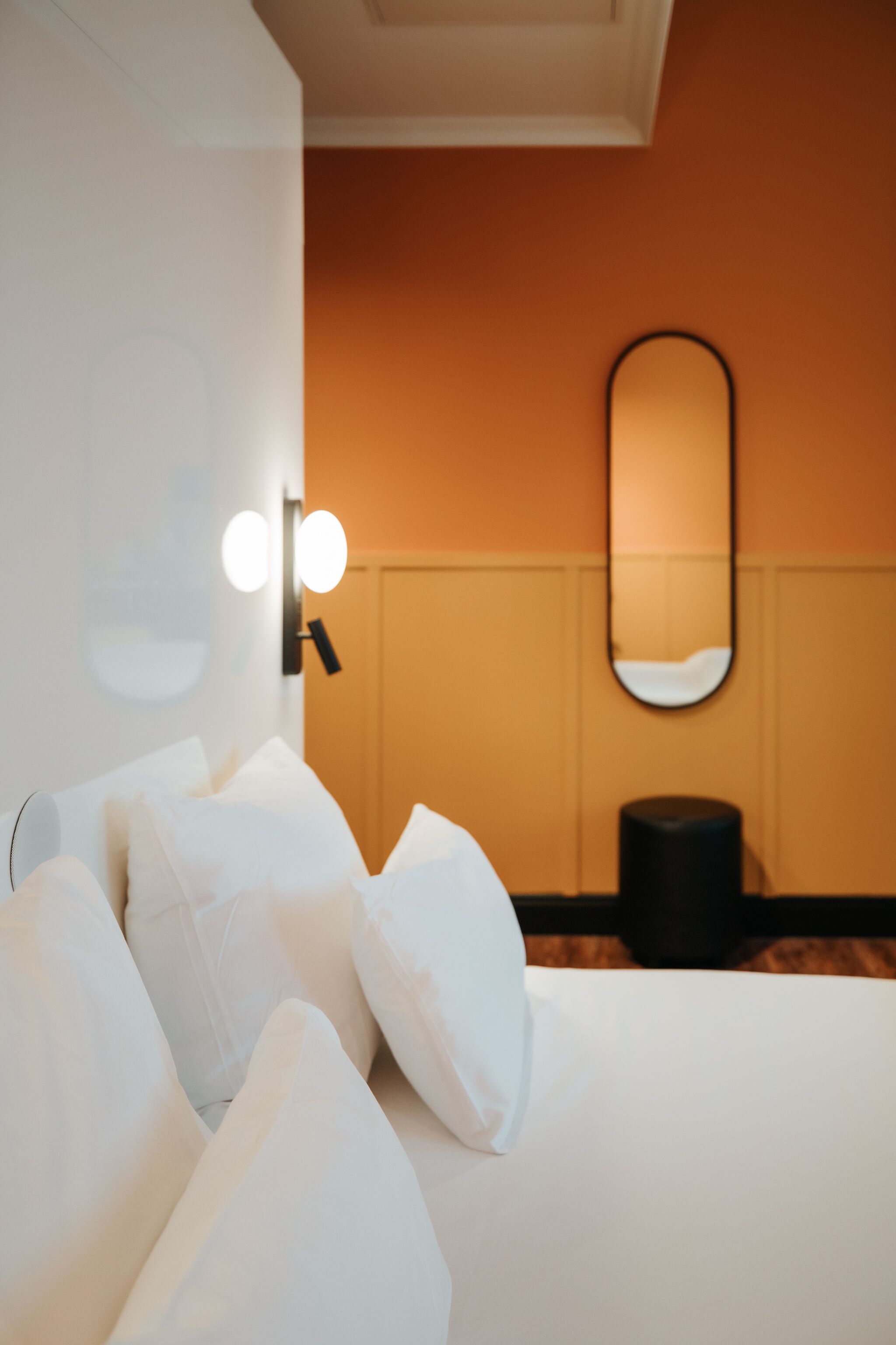 A hotel room with a bed, white pillows, a round mirror, and a wall-mounted light fixture.