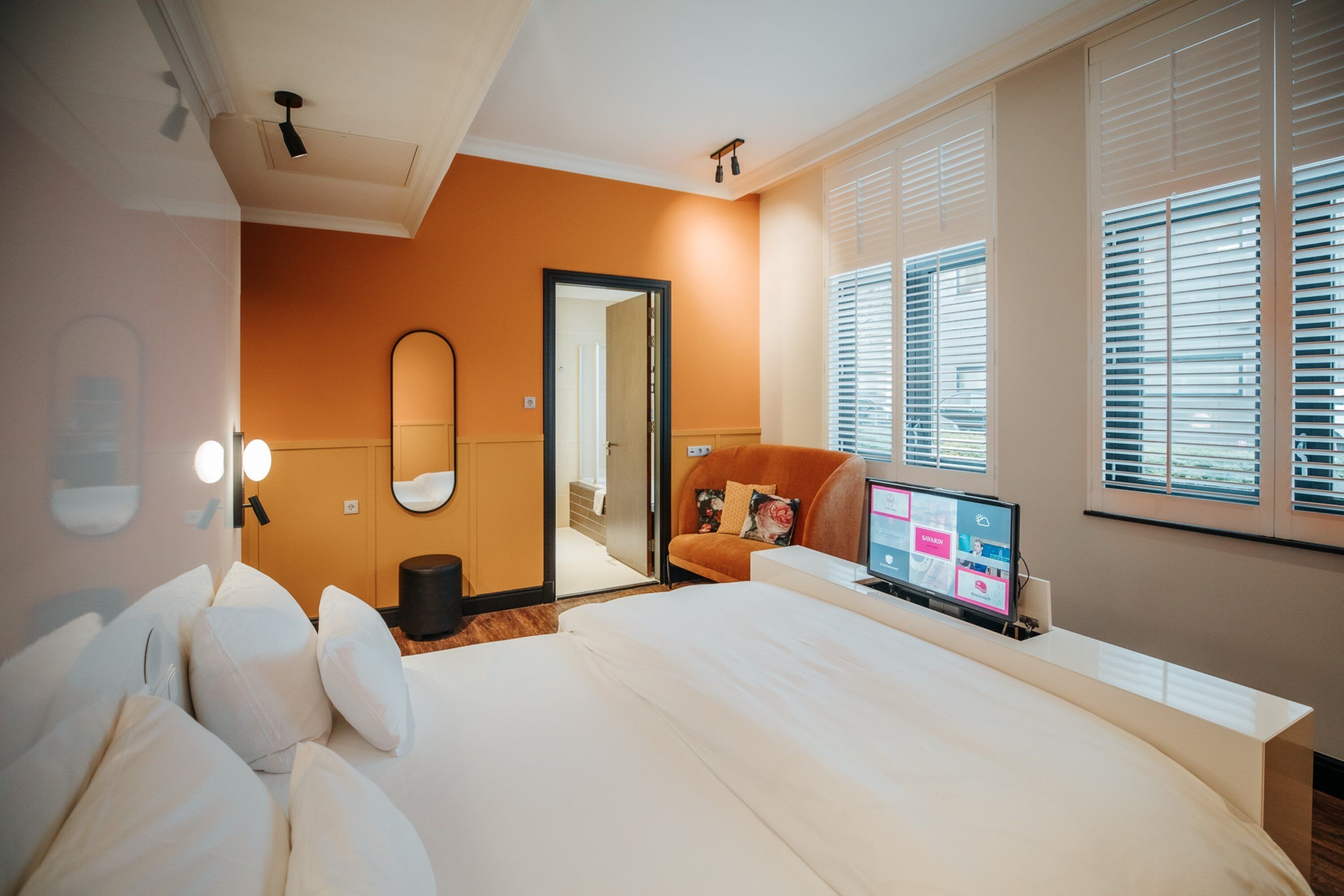 A modern hotel room with a large bed, a desk with a computer, and a window with blinds.