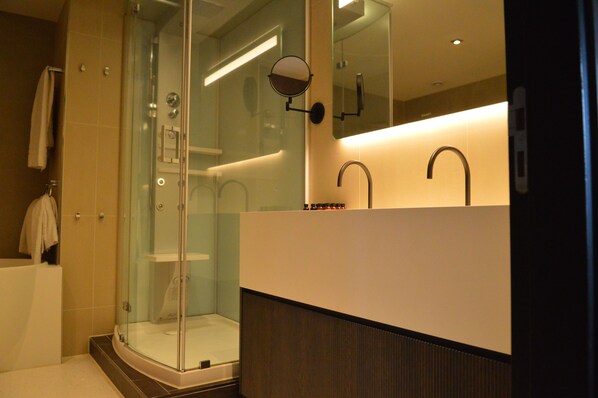 A modern bathroom with a glass-enclosed shower, a large mirror, and a white vanity with a dark wood panel.