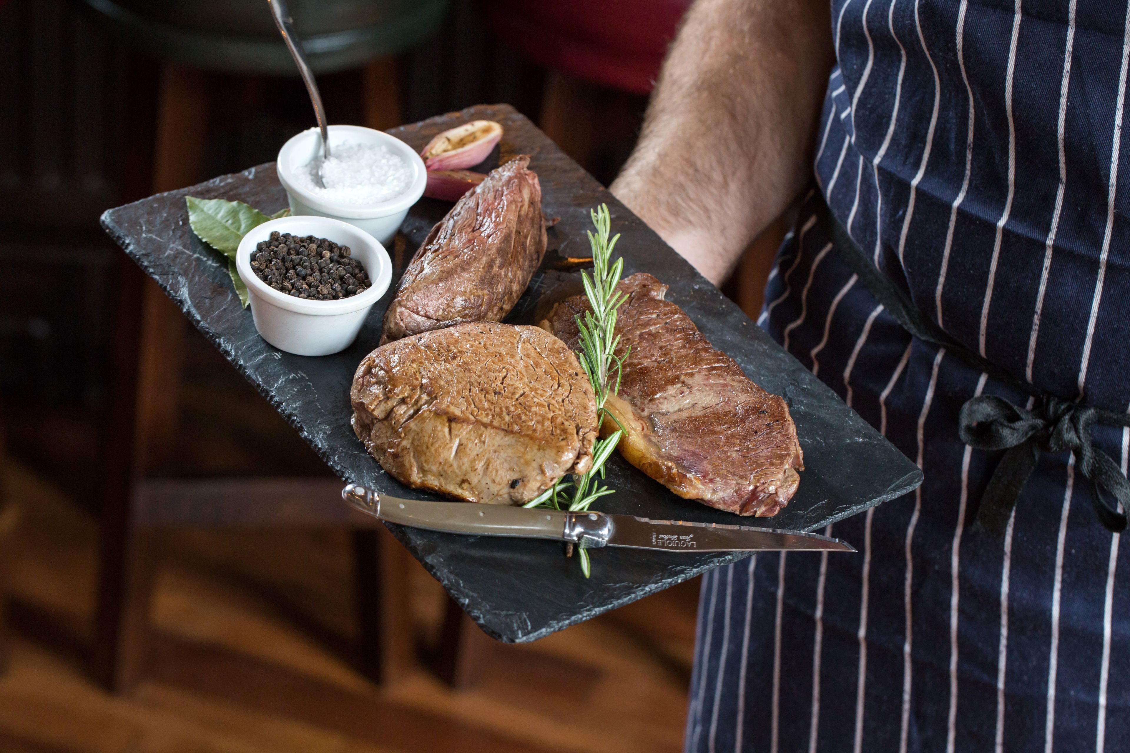 A person holding a black slate board with two pieces of meat, a sprig of rosemary, and two small bowls containing black pepper and salt.