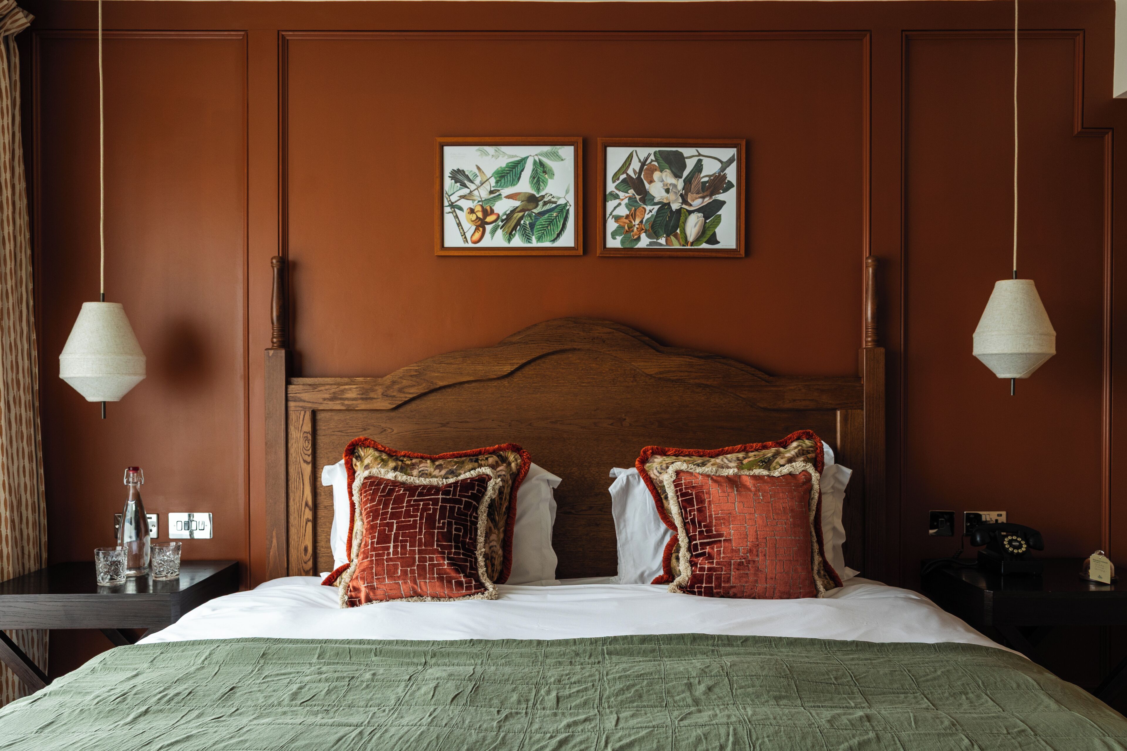 A hotel room with a wooden headboard, two framed botanical prints on the wall, a bed with red and white bedding, and two bedside tables with lamps.