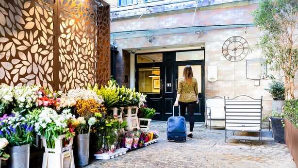 Entrada de hotel com diversas flores e plantas, um banco de metal e uma pessoa com mala.