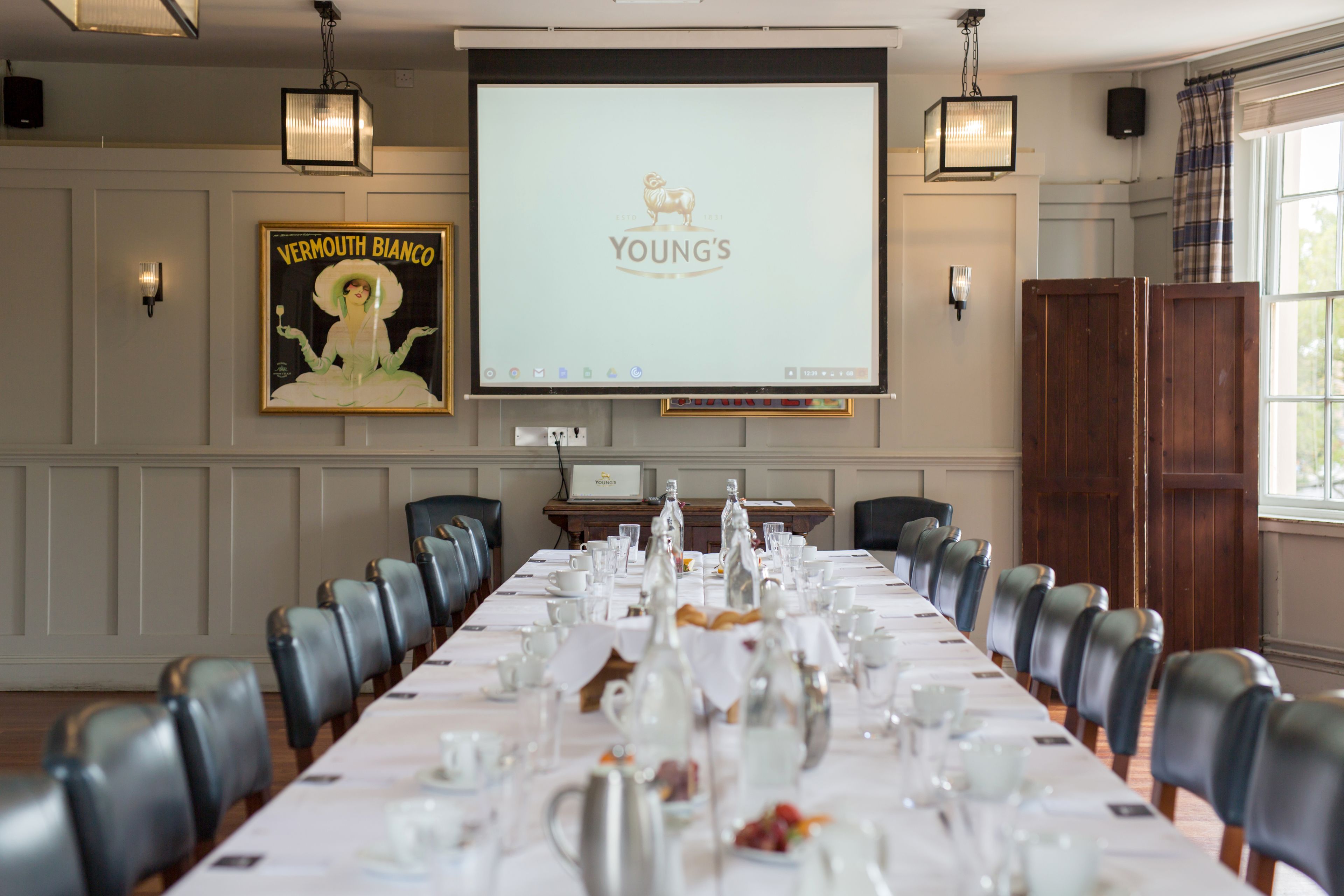 A conference room with a long table set for a meeting, a projector screen displaying 'YOUNG'S', and a poster of 'VERMOUTH BIANCO'.
