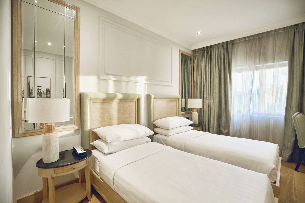 Une chambre d’hôtel avec deux lits, une table de chevet avec une lampe, un miroir et une fenêtre avec des rideaux.