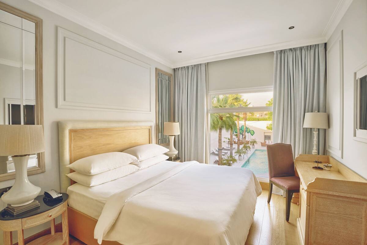 Une chambre d’hôtel avec un grand lit, un bureau, une chaise, une lampe et une vue sur une piscine et des palmiers.