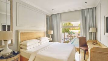 Habitación de hotel con una cama grande, un escritorio, una silla, una lámpara y vista a una piscina y palmeras.