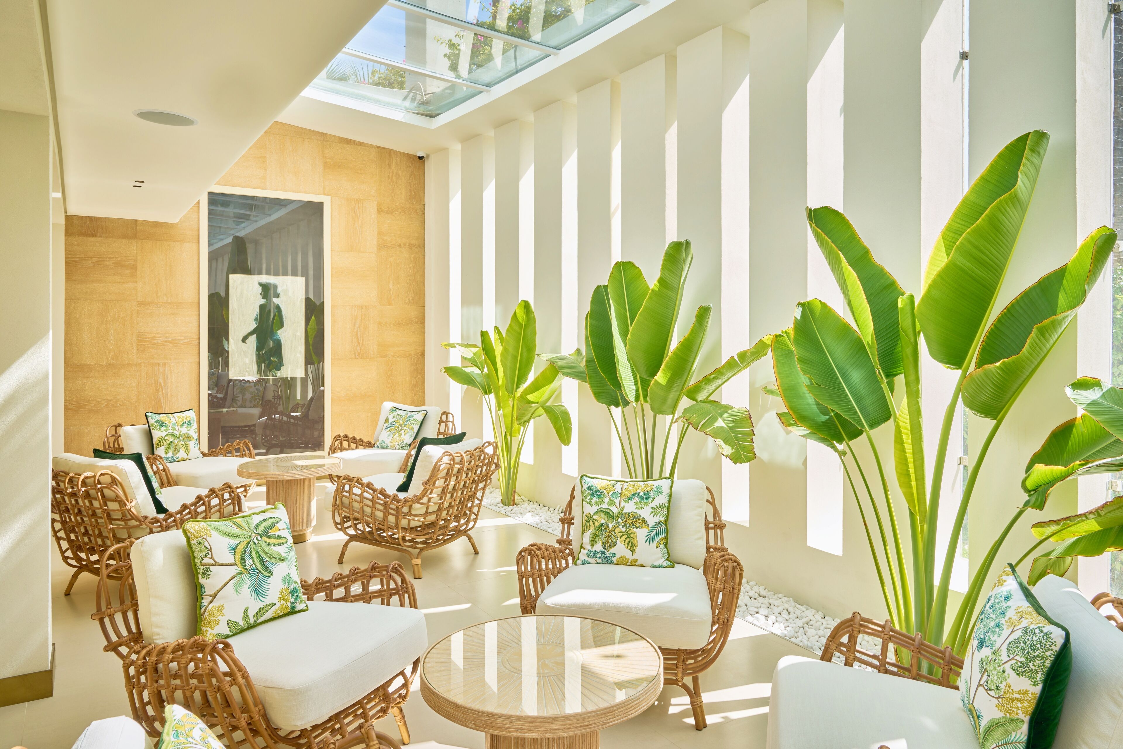 Un salón luminoso e inundado de sol, con mobiliario de mimbre, plantas verdes grandes y techo de cristal.