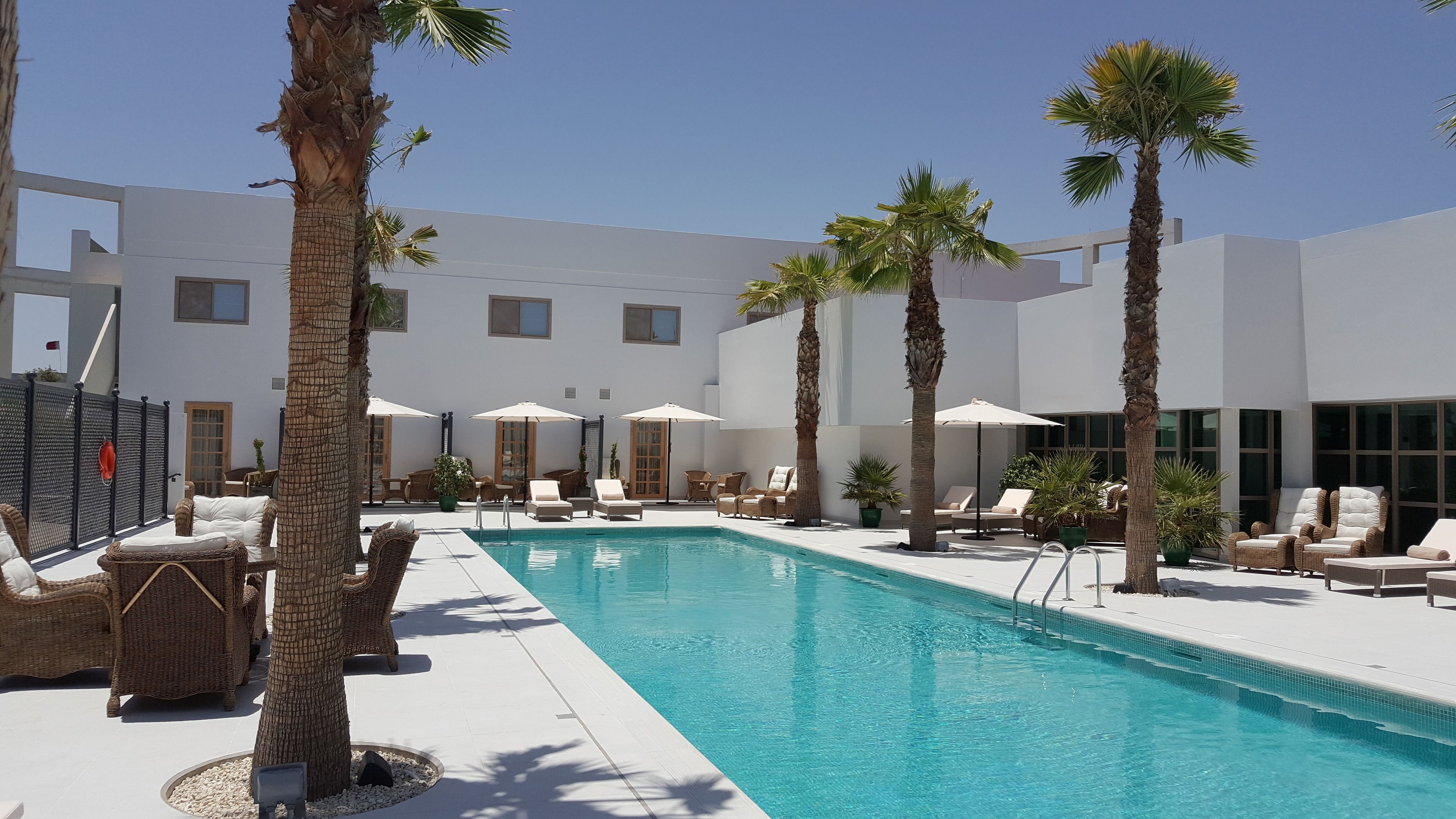 A pool area with lounge chairs and umbrellas, surrounded by palm trees and a modern building.