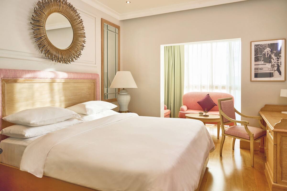 Une chambre d’hôtel avec un grand lit, un bureau, une chaise, une lampe, un miroir et une fenêtre avec des rideaux.