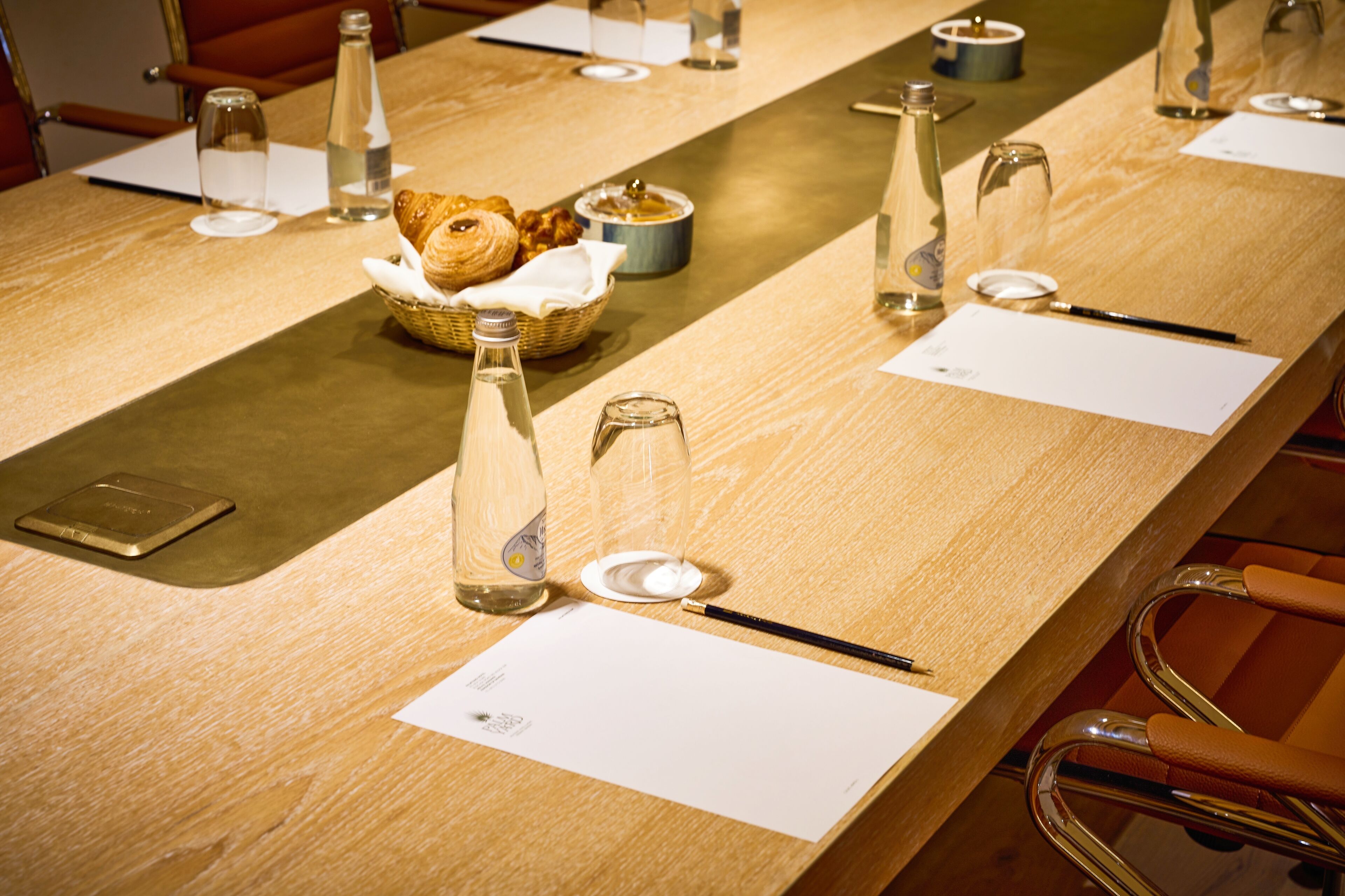 A conference table with documents, a basket of pastries, and glass bottles.
