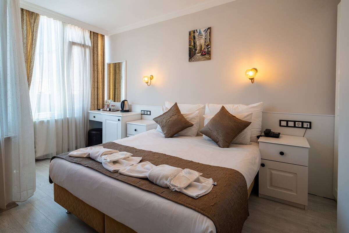 A hotel room with a bed, bedside tables, a desk, and a painting on the wall.