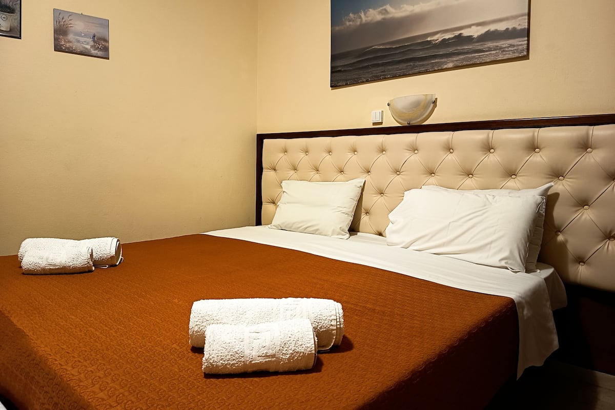A neatly made bed with white linens and two towels, a tufted headboard, and a painting of a seascape on the wall.