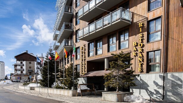 A multi-story building with a restaurant named "Ristorante Banchette" on the ground floor. There are flags, a signboard, and a small outdoor seating area with a table and chairs. - Hotel Cristallo (Sestriere)